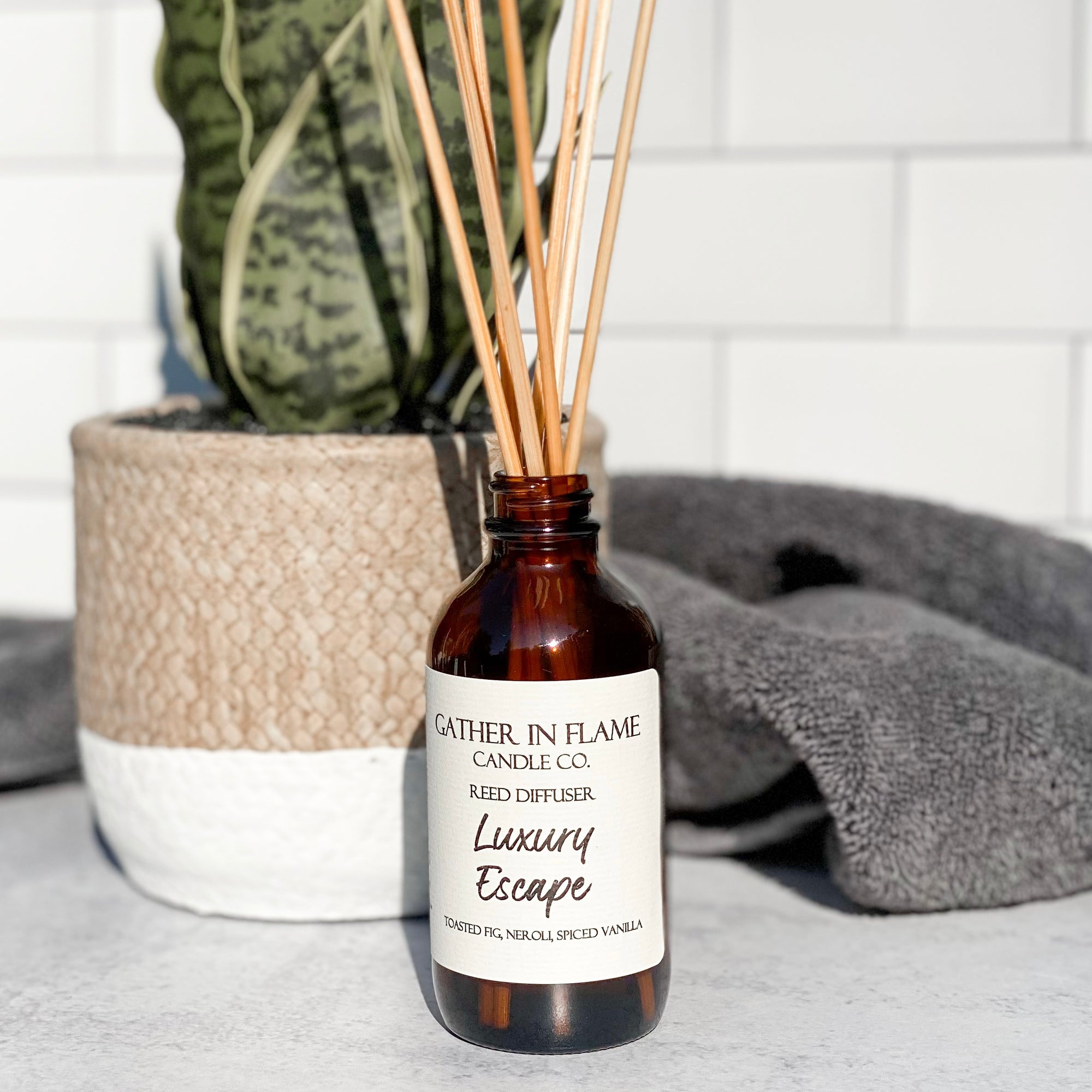 A brown glass reed diffuser bottle with a label reading "Luxury Escape", placed on a grey surface, surrounded by plant and towels with white tile background.