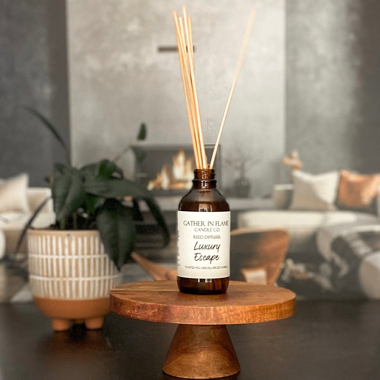 A brown glass reed diffuser bottle with a label reading "Luxury Escape", placed on a wood surface, with a plant and living room in the background.