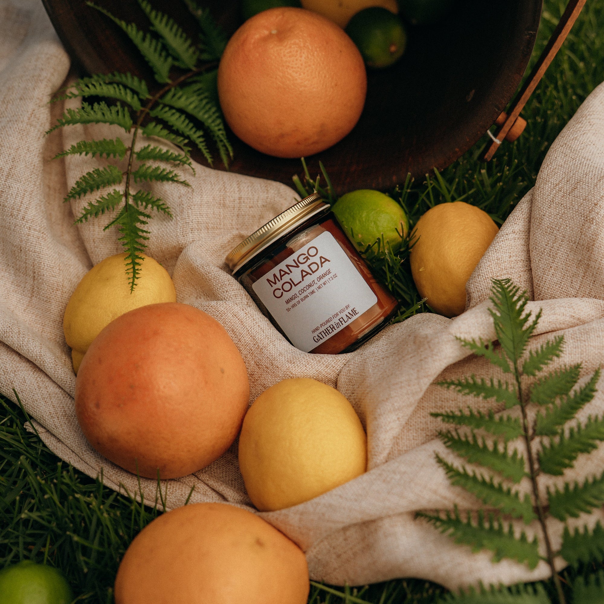 Mango Colada jar surrounded by mangoes and limes on a natural fabric with greenery.