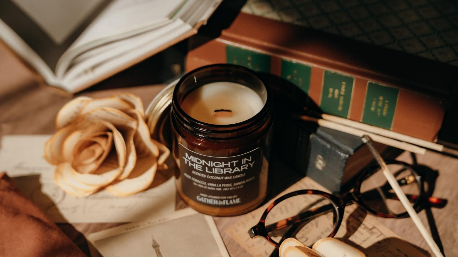Amber glass jar candle labeled Midnight in the Library surrounded by books, glasses and ephemera