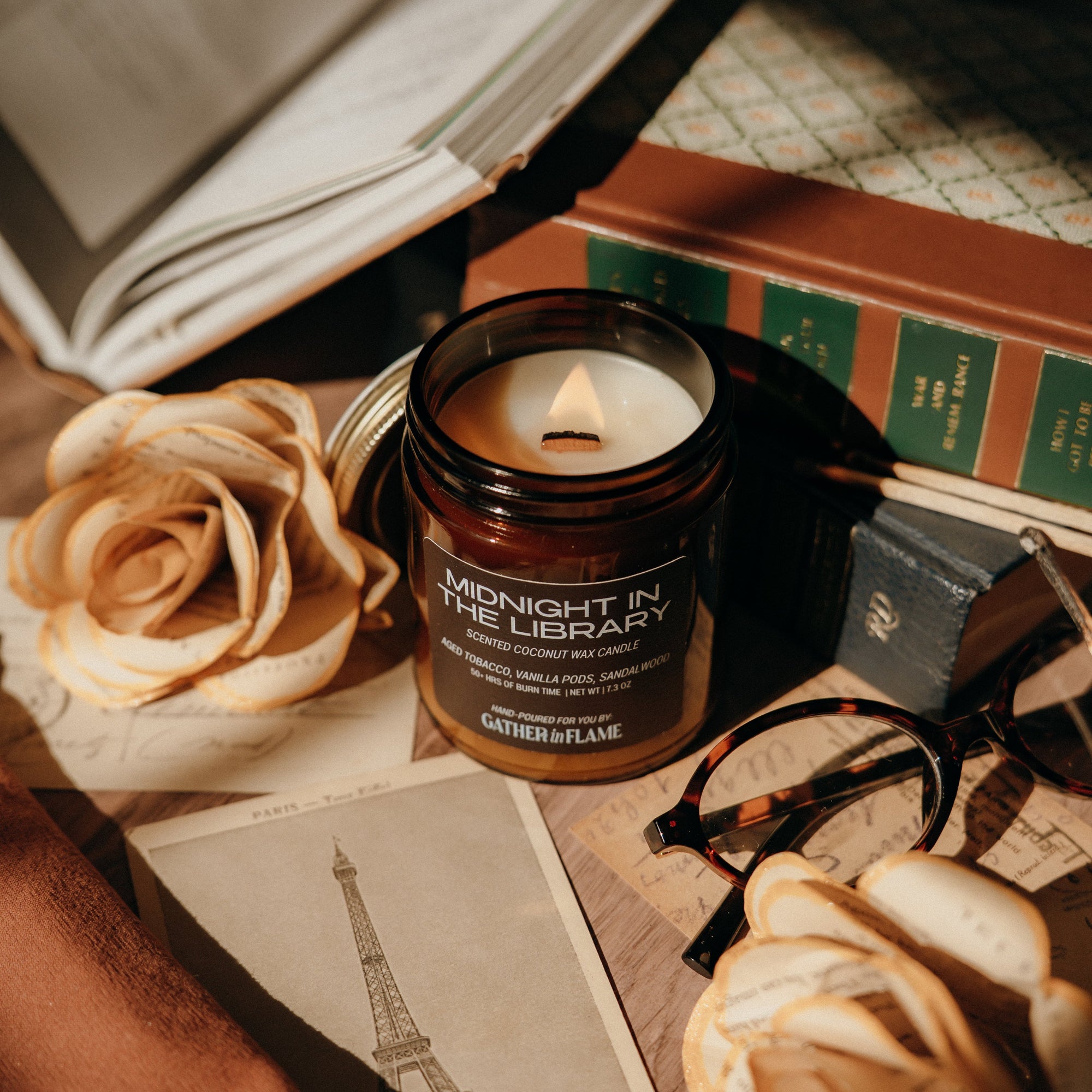 Candle in a jar labeled 'Midnight in the Library' on a table with books, roses, and glasses.