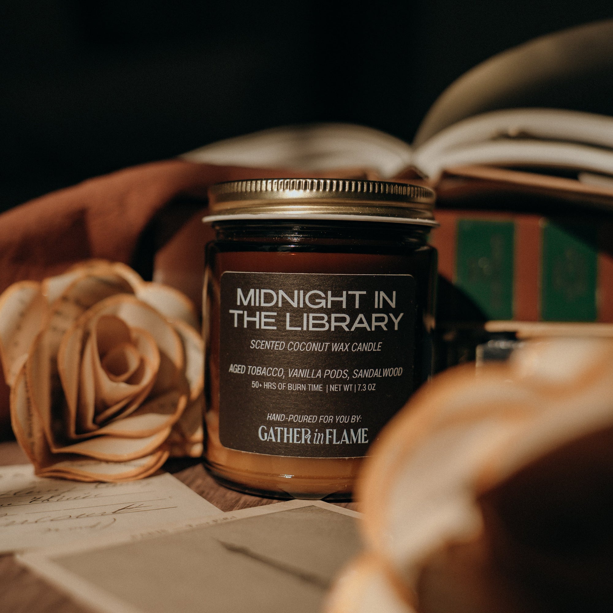 Candle jar labeled 'Midnight in the Library' by Gather of Flame on a surface with a rose and books.
