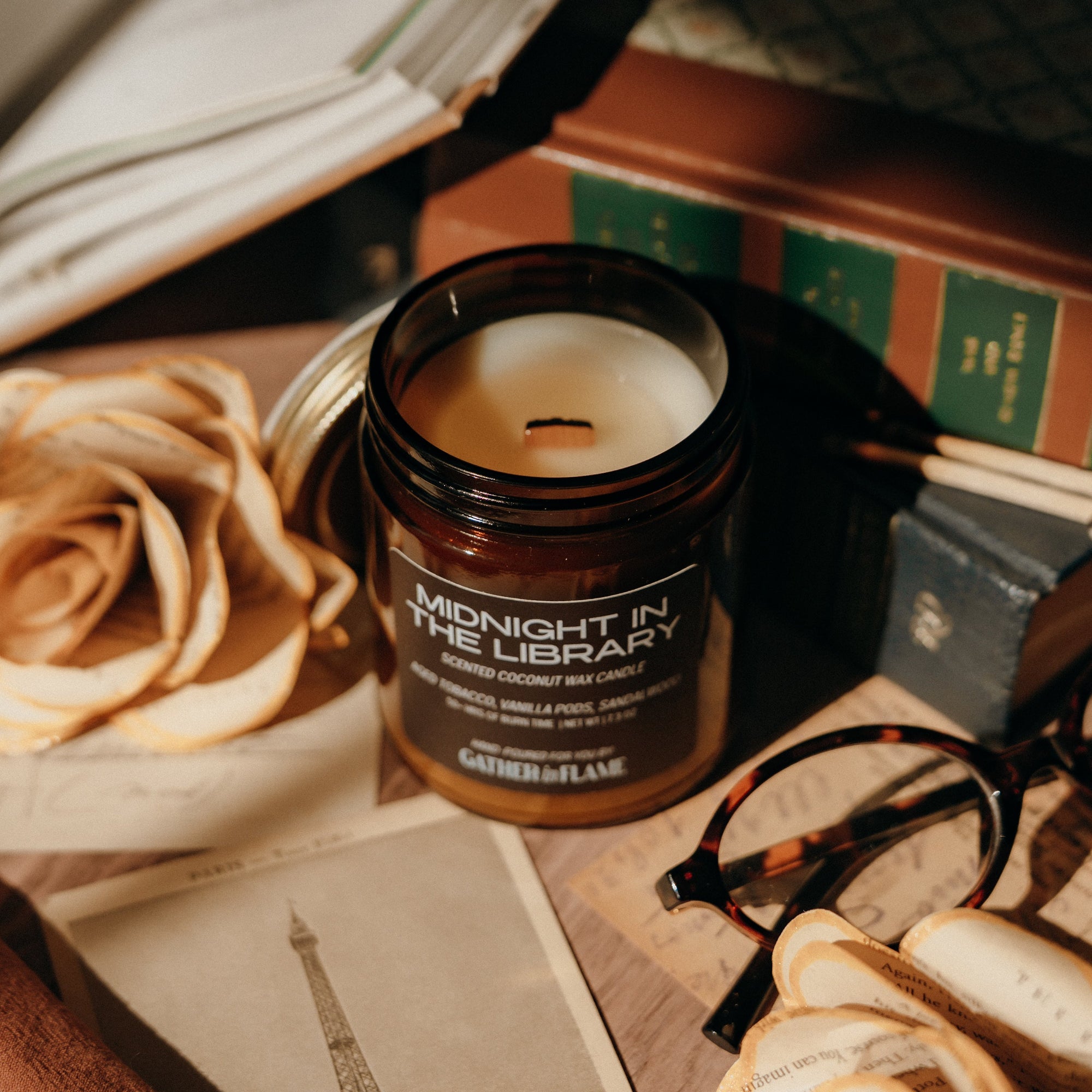 Jar of candle labeled 'Midnight in the Library' on a table with books and flowers