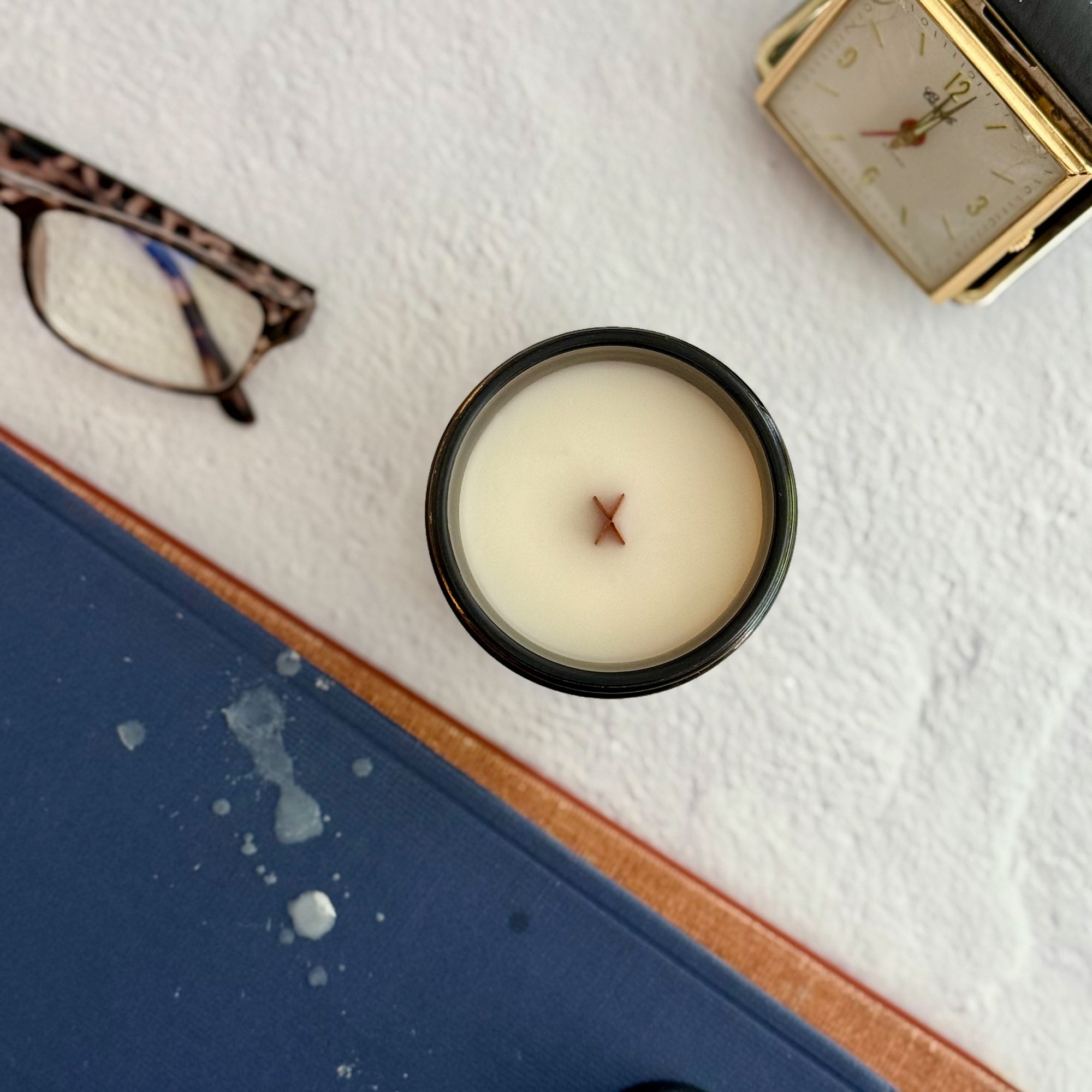 Topdown view of candle with wood wick on a textured surface with glasses, books and a clock in the background.