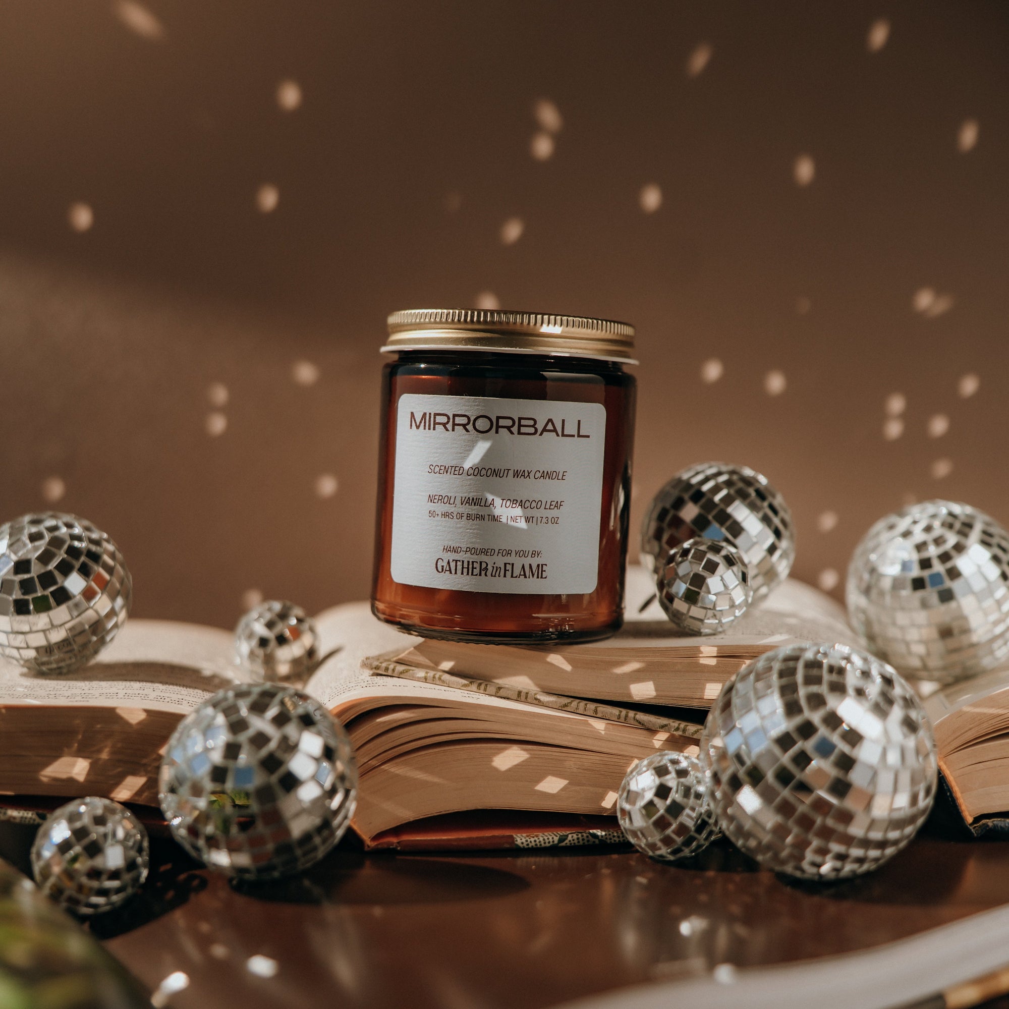 Jar of 'Mirrorball' candle on an open book with disco balls around it, on a dark background.