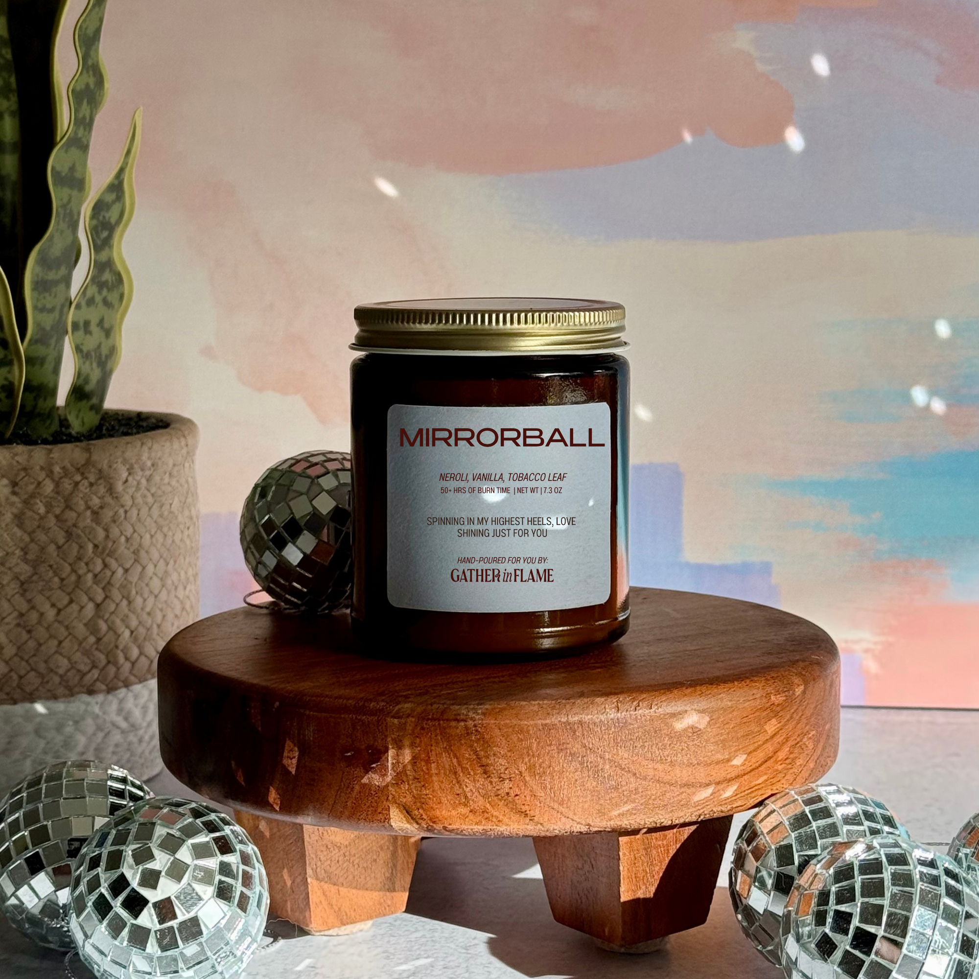 Candle jar labeled 'Mirrorball' on a wooden stand with decorative disco balls and a plant in the background.