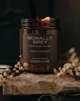 Candle labeled 'Morally Grey' by Gathered Flame on a dark background with dried flowers and scissors.