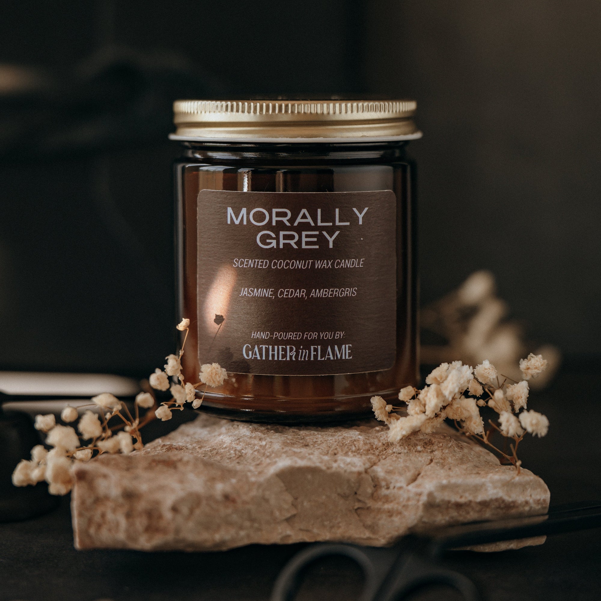 Candle jar labeled 'Morally Grey' by Gather the Flame on a stone surface with flowers and scissors.