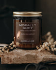 Candle jar labeled 'Morally Grey' by Gather the Flame on a stone surface with flowers and scissors.