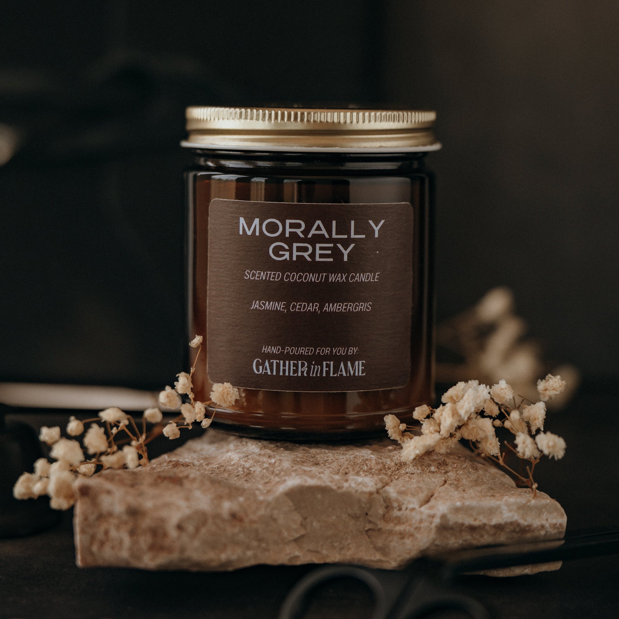 Candle labeled 'Morally Grey' by Gather of Flame on a stone surface with flowers and scissors.