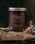 Candle labeled 'Morally Grey' by Gather of Flame on a stone surface with flowers and scissors.