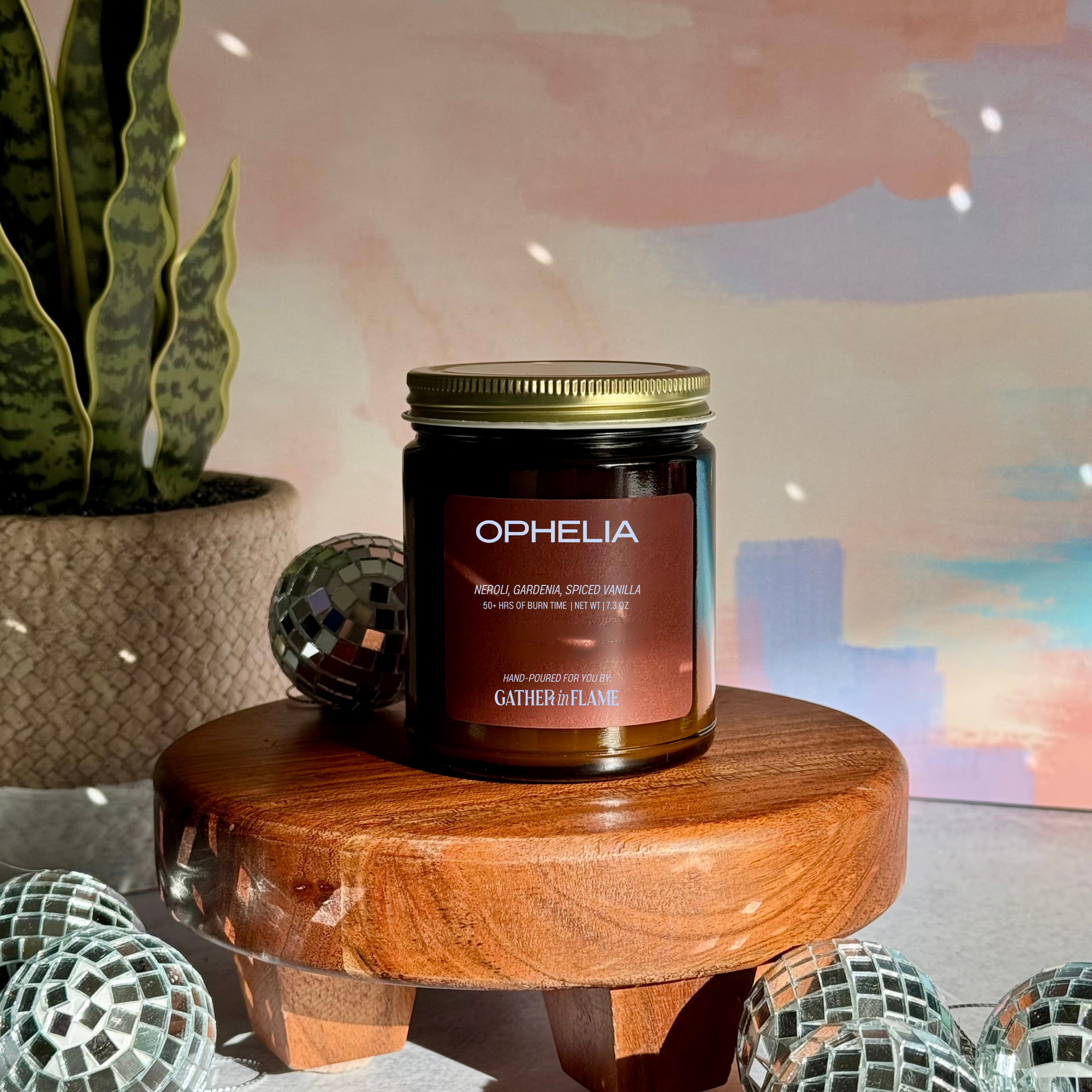 Candle jar labeled 'Ophelia' on a wooden surface with disco balls and a plant in the background.