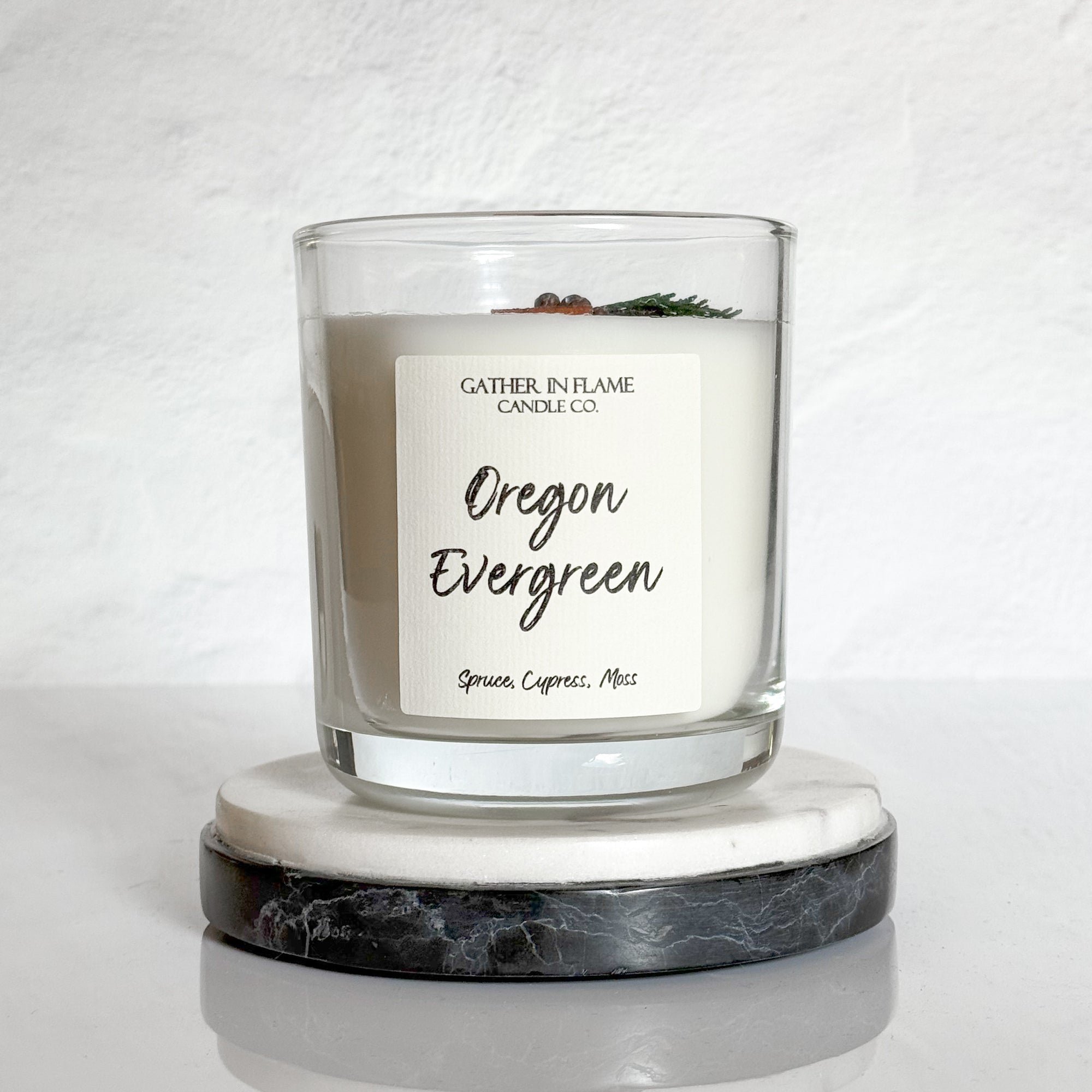 Closeup of clear glass 12oz Oregon Evergreen candle with wood wick and juniper sprig on surface