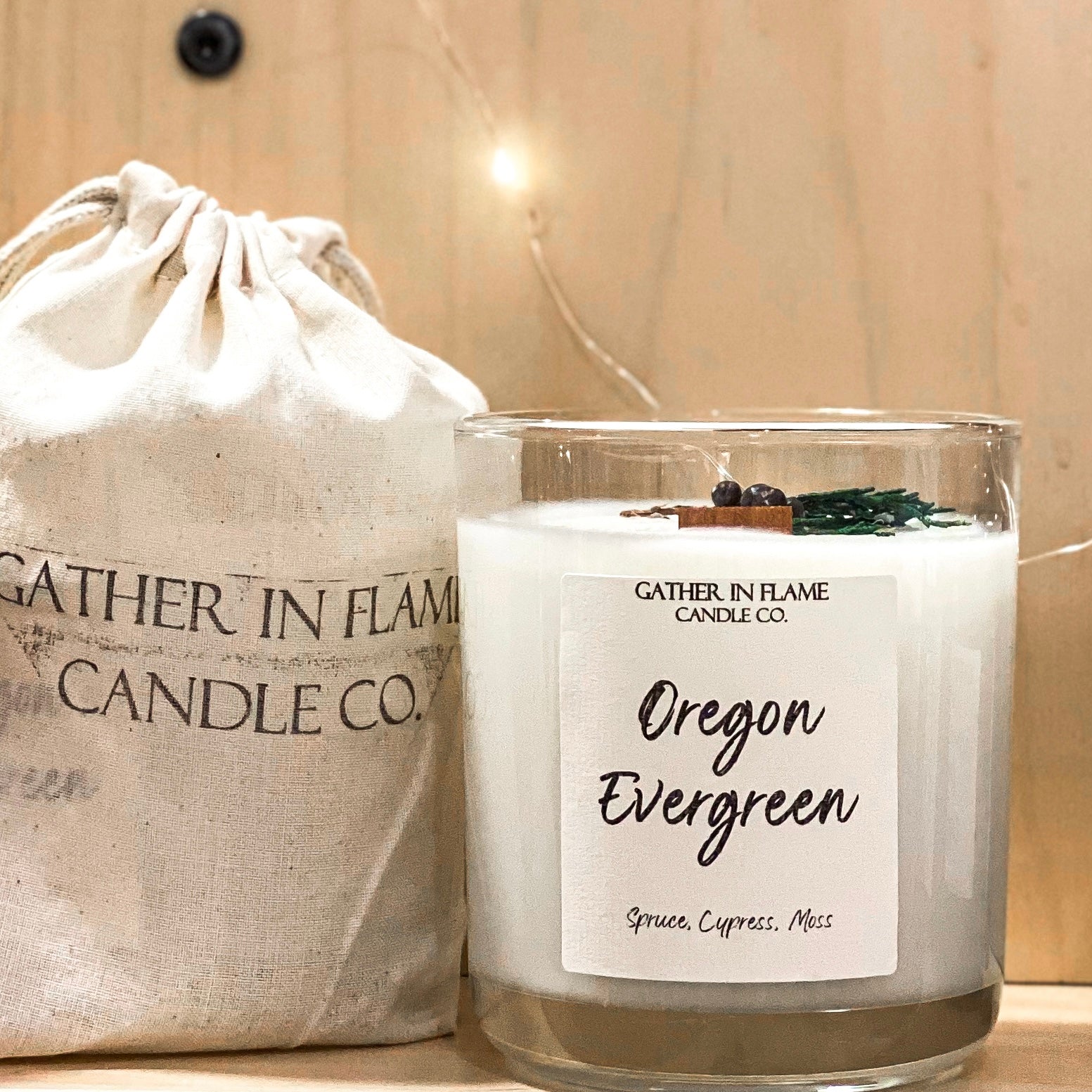 Clear glass 12oz Oregon Evergreen candle  on a shelf next to a muslin bag labeled Gather in Flame Candle Co
