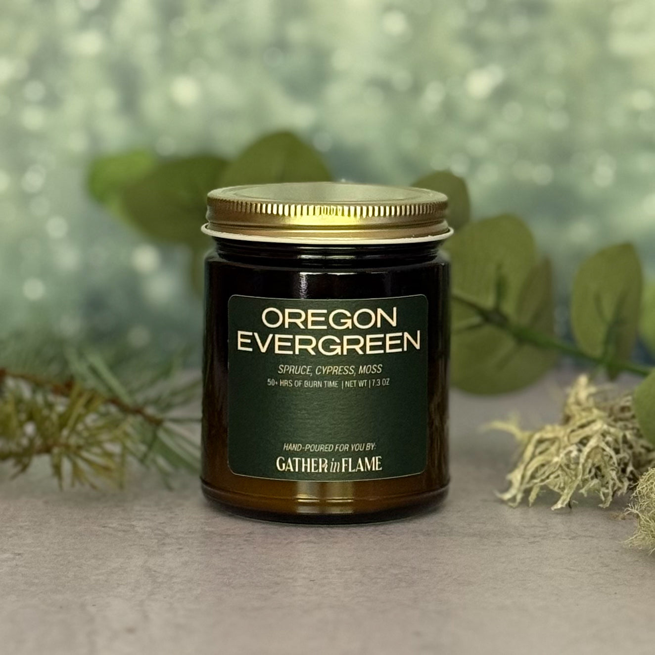 Jar of 'Oregon Evergreen' candle with a gold lid on a grey surface with pine branches, moss and eucalyptus