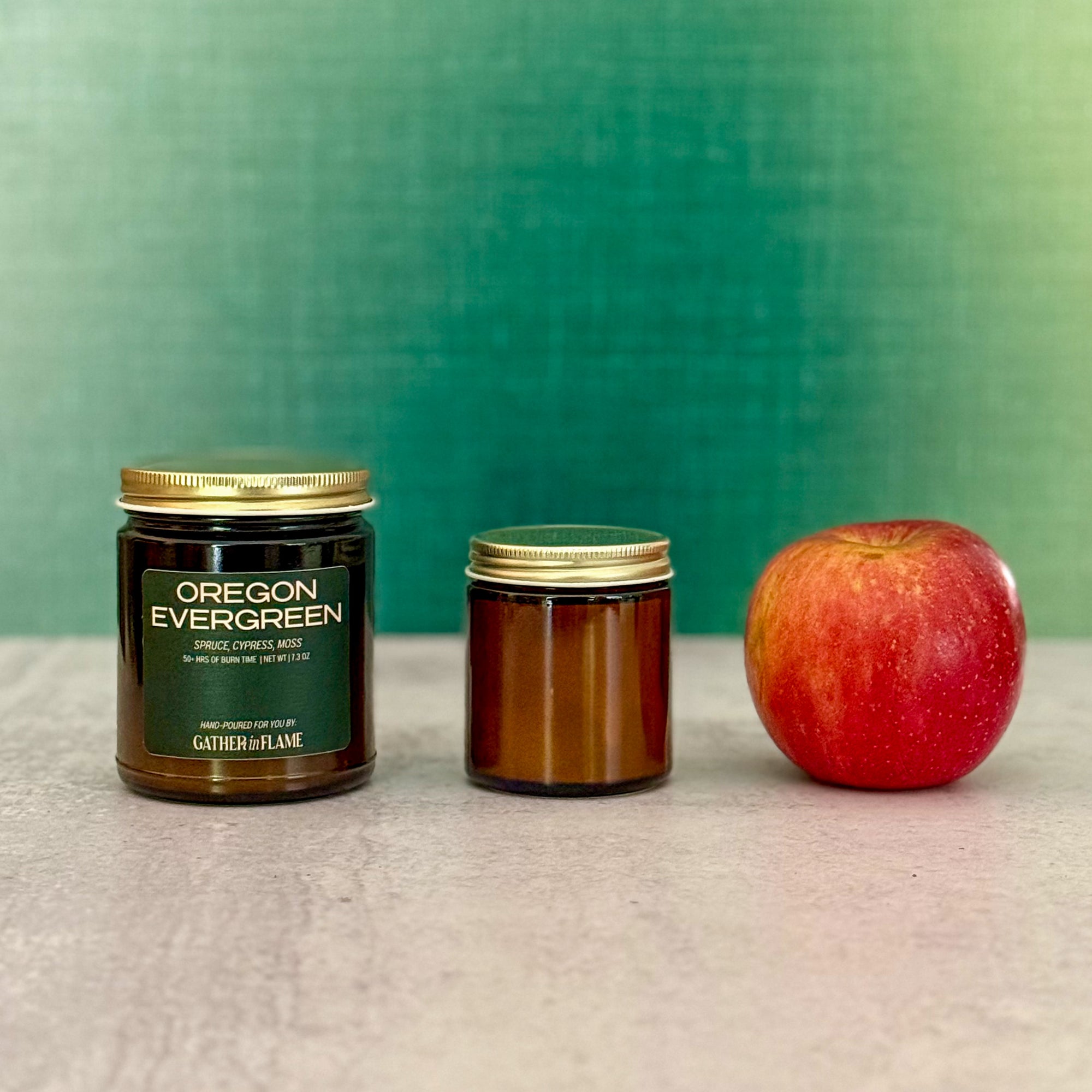 Two candles of different sizes labeled Oregon Evergreen next to an apple with a green background