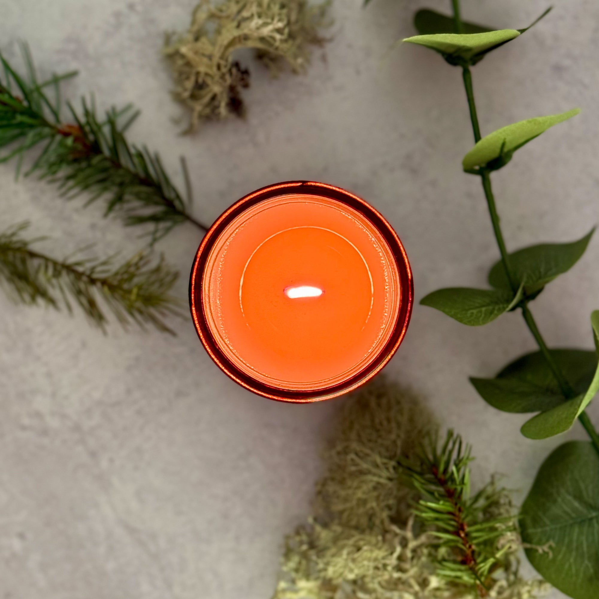 Overhead view of lit 'Oregon Evergreen' candle on a grey surface with pine branches, moss and eucalyptus