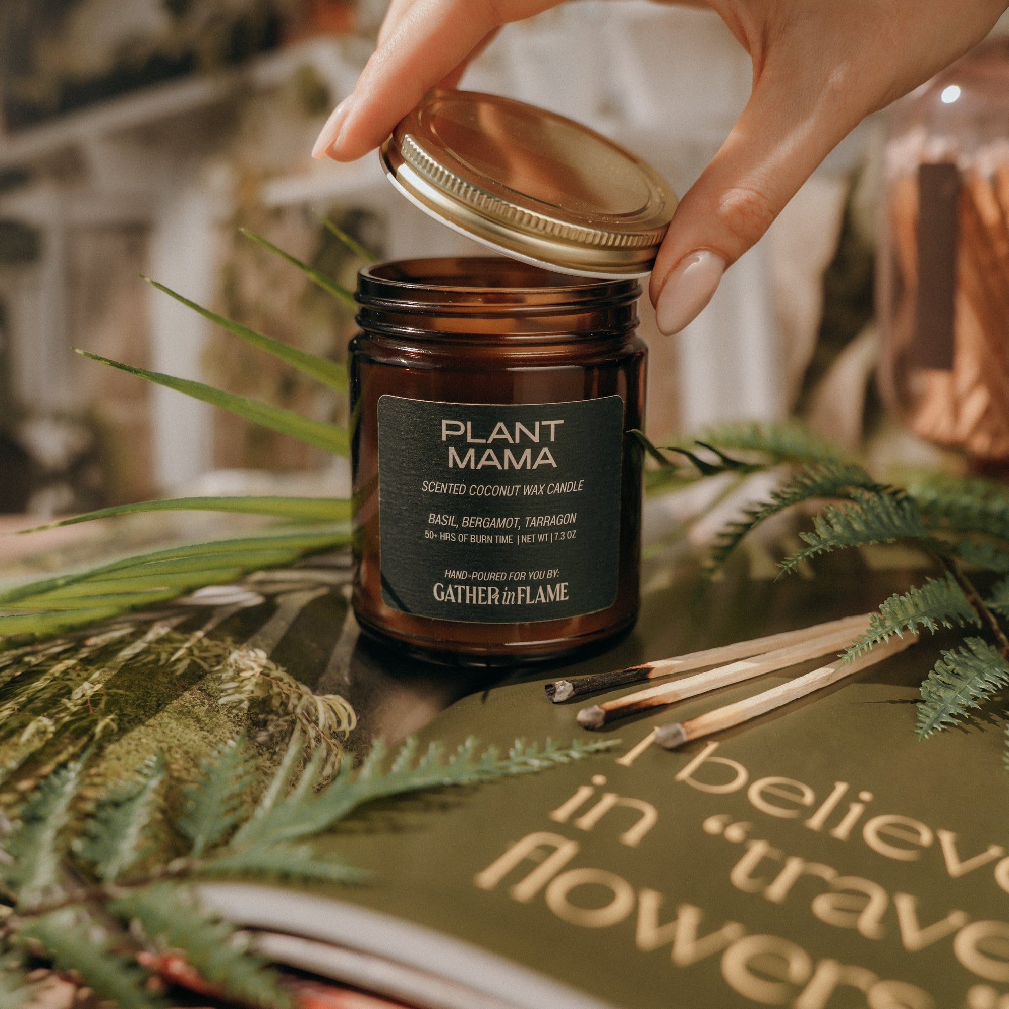 Hand opening a 'Plant Mama' candle jar with greenery and a quote in the background