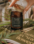 Hand opening a 'Plant Mama' candle jar with greenery and a quote in the background
