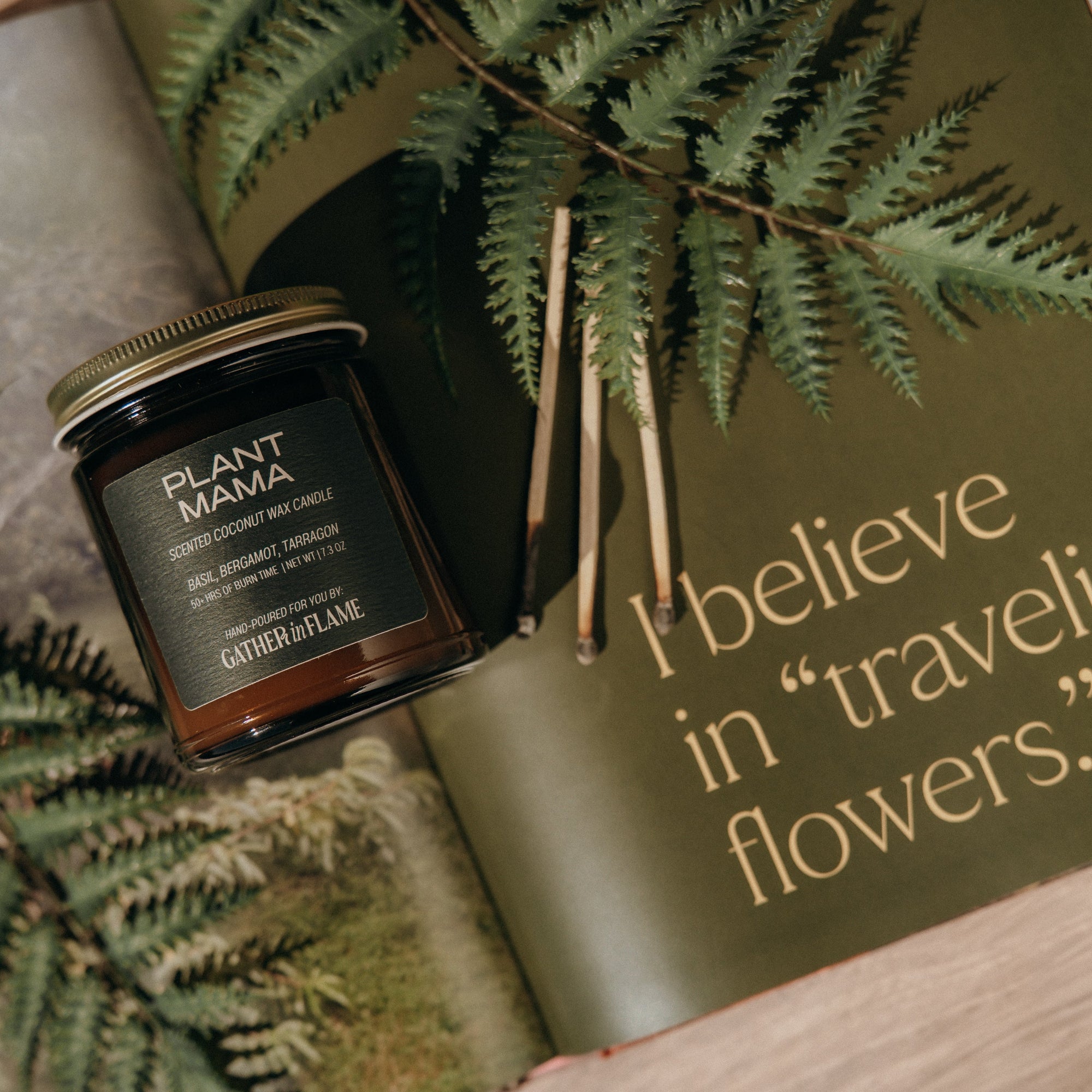 Jar of candle labeled 'Plant Mama' on a book with text 'I believe in traveling flowers.' surrounded by fern leaves.