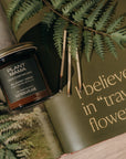 Jar of candle labeled 'Plant Mama' on a book with text 'I believe in traveling flowers.' surrounded by fern leaves.