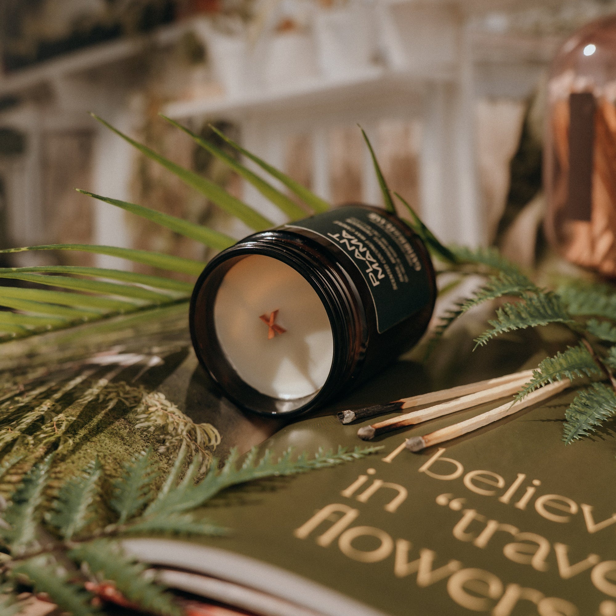 Candle with a decorative lid on a surface with greenery and a book in the background