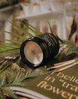 Candle with a decorative lid on a surface with greenery and a book in the background