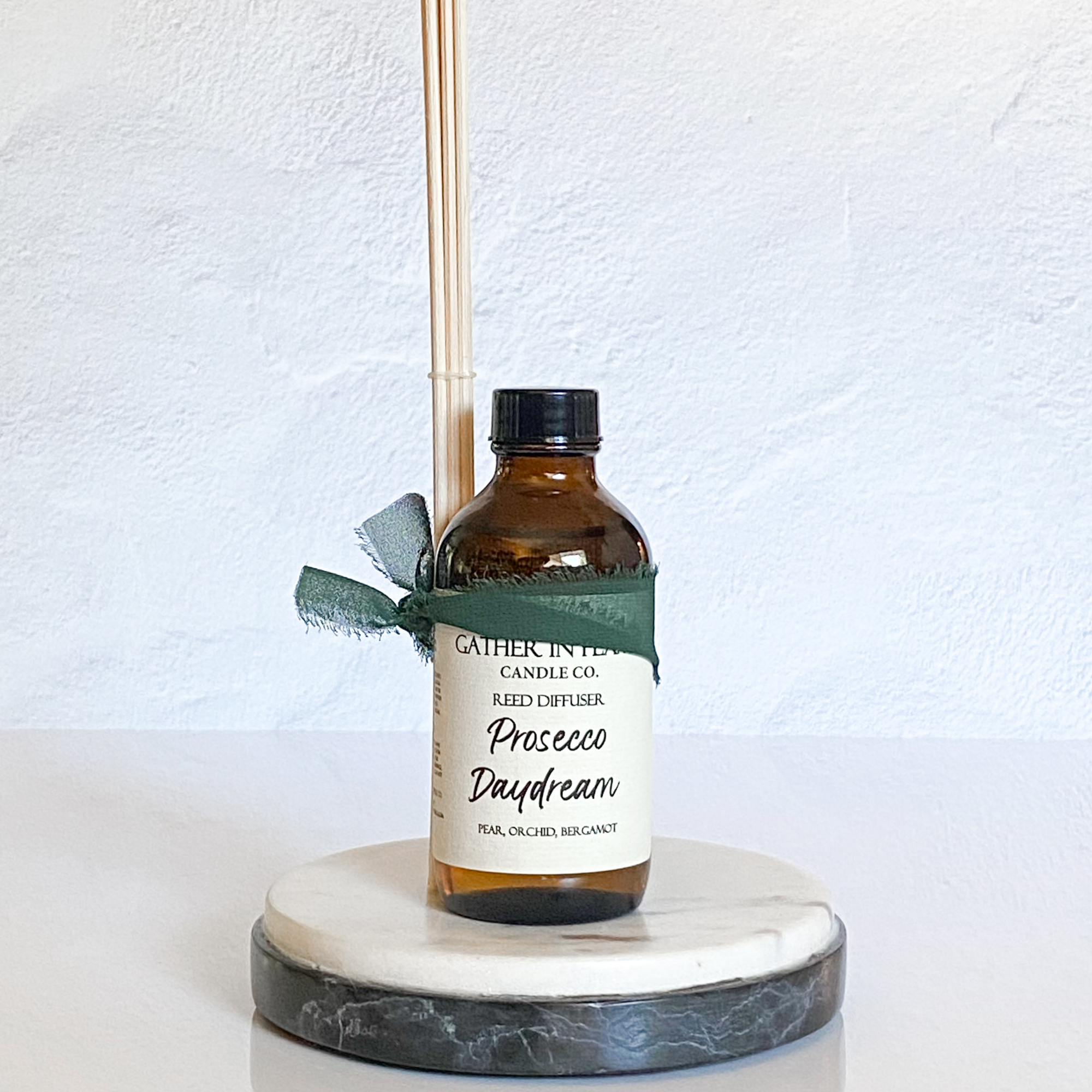 A Reed diffuser with an amber glass bottle. There is a green ribbon and a label with the product name 'Prosecco Daydream' written on it placed on a marble stand.