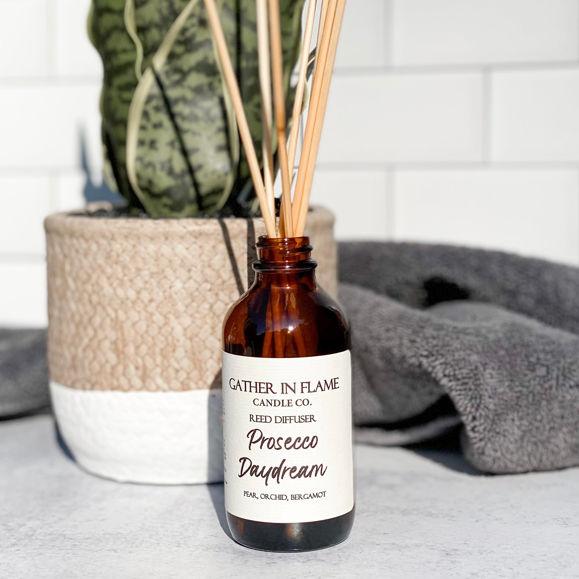 A brown glass reed diffuser bottle with a label reading "Prosecco Daydream", placed on a grey surface, surrounded by plant and towels with white tile background.