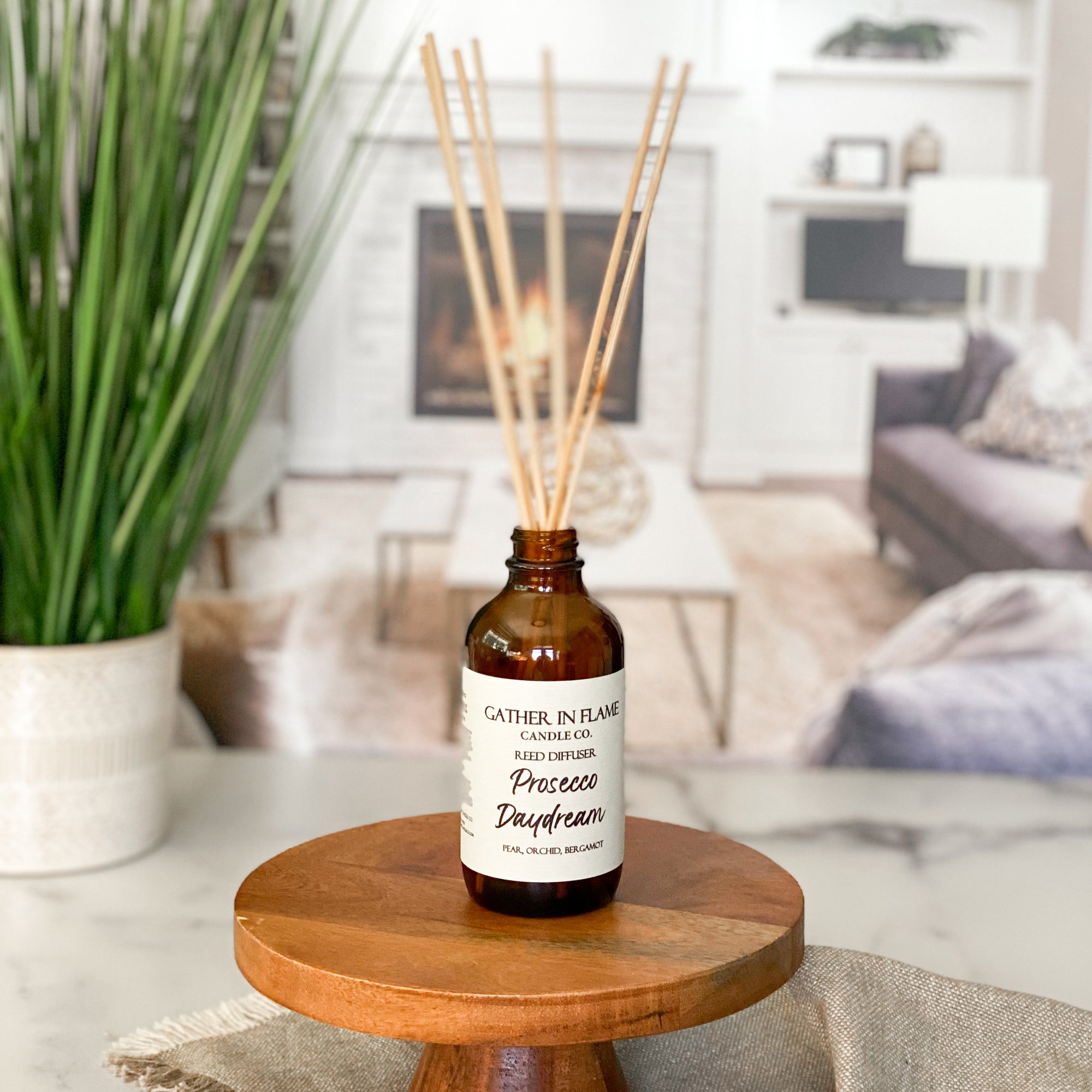 A brown glass reed diffuser bottle with a label reading "Prosecco Daydream", placed on a wood surface, with a plant and living room in the background
