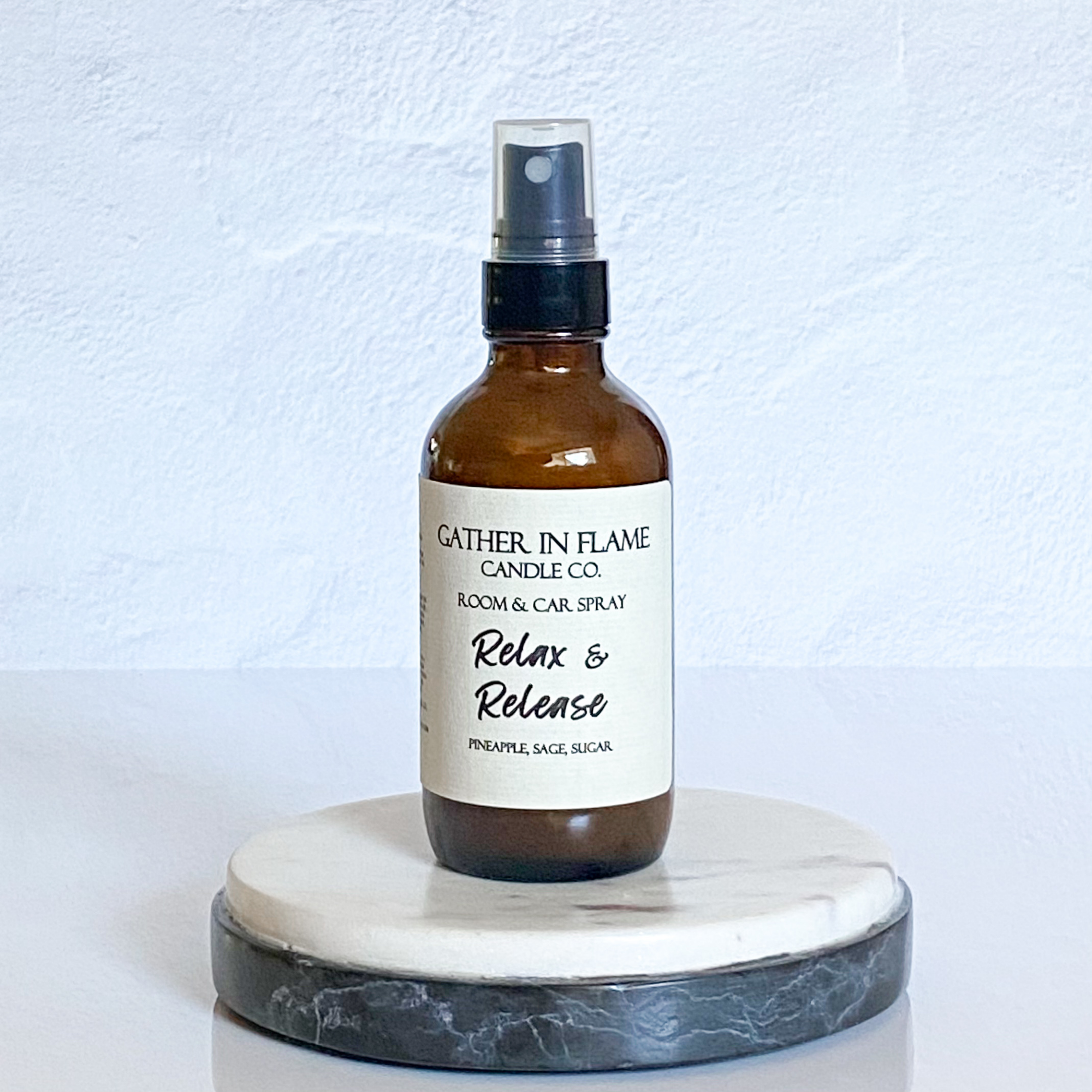 A brown bottle with a white label that reads 'GATHER IN FLAME CANDLE CO. ROOM & CAR SPRAY Relax & Release Pineapple Sage' on a white marble surface.