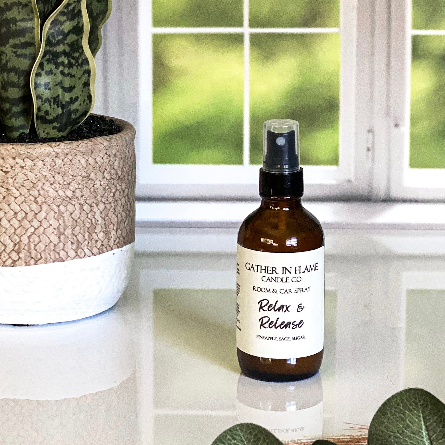 A brown amber spray bottle labeled 'Relax & Release' by Gather In Flame Candle Co. on a white marble surface with a window and plant in the background.