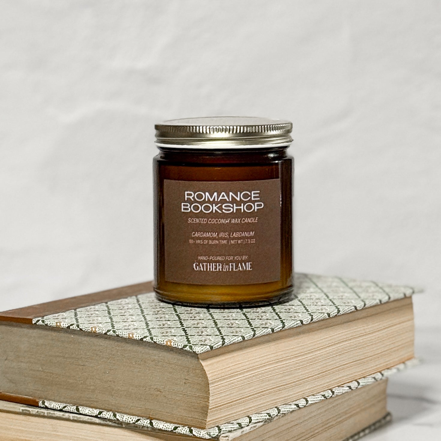 Amber glass jar candle labeled Romance Bookshop on top of a stack of books with a white background.