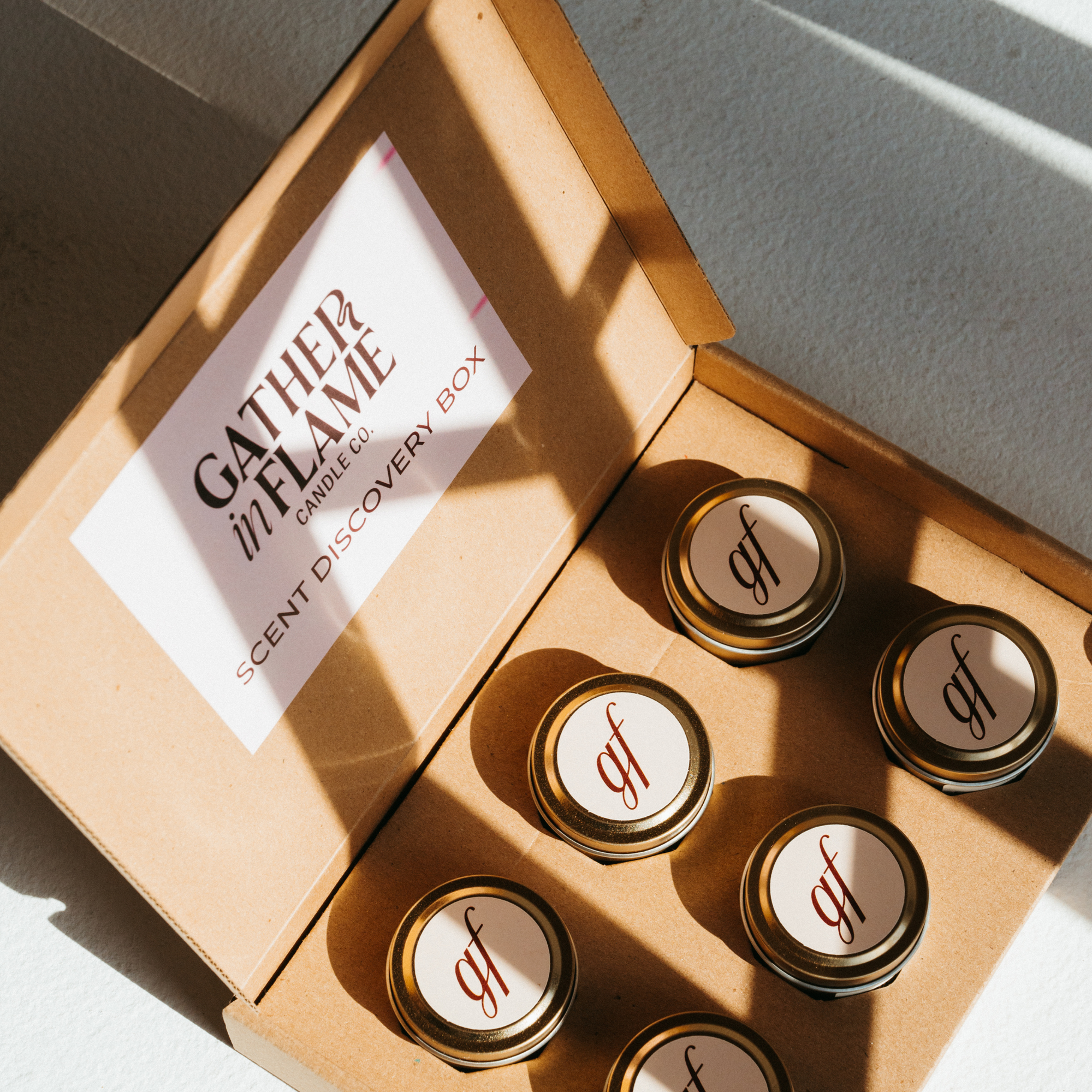 An open cardboard box labeled 'GATHER IN FLAME' with six small coconut wax candles arranged inside, each with a wooden wick.