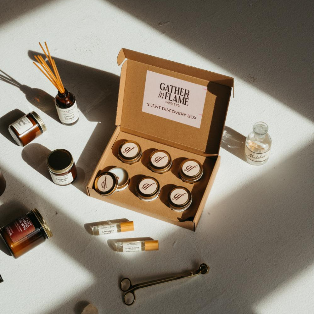 Scent discovery box surrounded by candles, reed diffusers, perfume oil rollers and match bottle on a white surface.