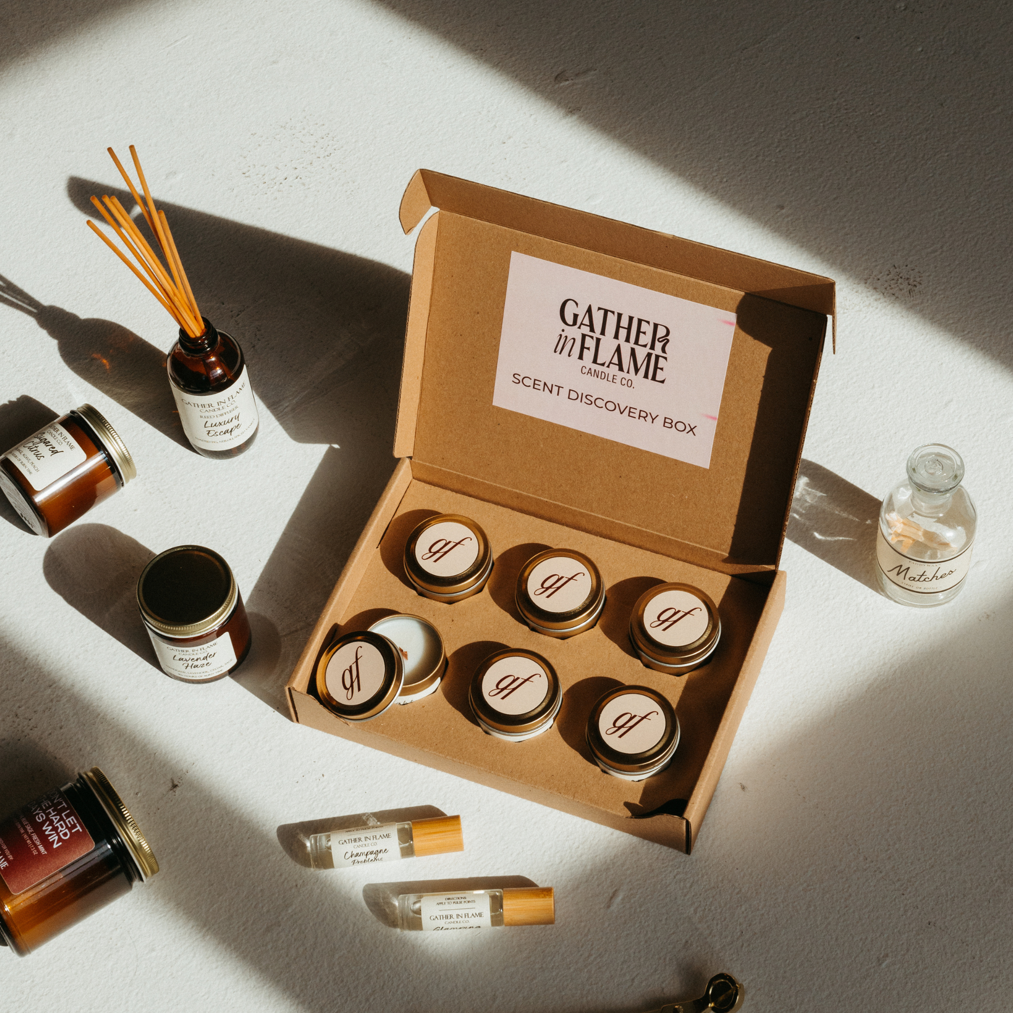 Scent discovery box surrounded by candles, reed diffusers, perfume oil rollers and match bottle on a white surface.