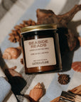 Amber glass candle jar labeled Seaside Reads on a towel surrounded by shells and driftwood.