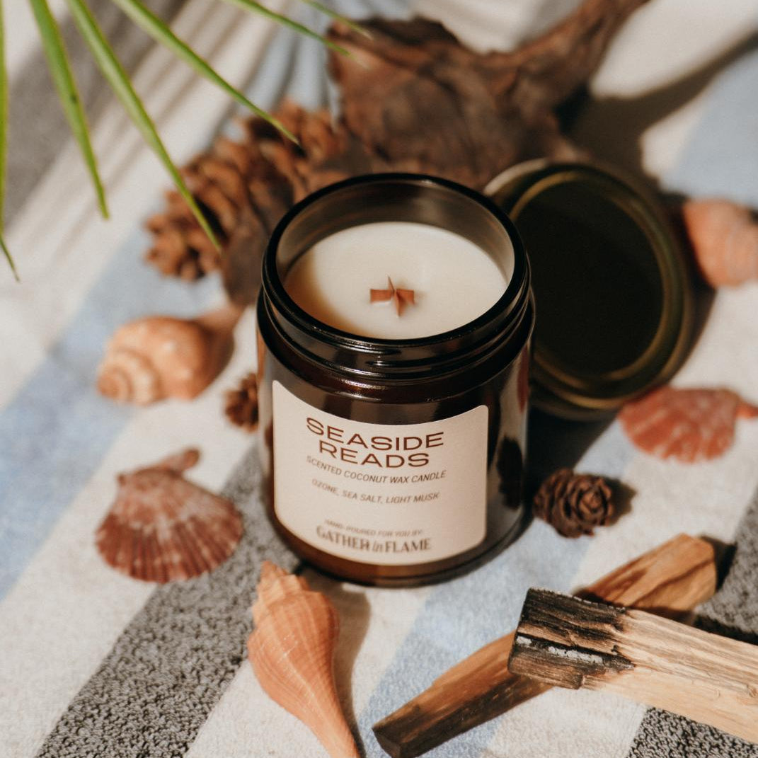 Amber glass candle jar labeled Seaside Reads with the lid off and candle top showing on a towel surrounded by shells and driftwood.