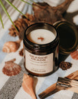Amber glass candle jar labeled Seaside Reads with the lid off and candle top showing on a towel surrounded by shells and driftwood.