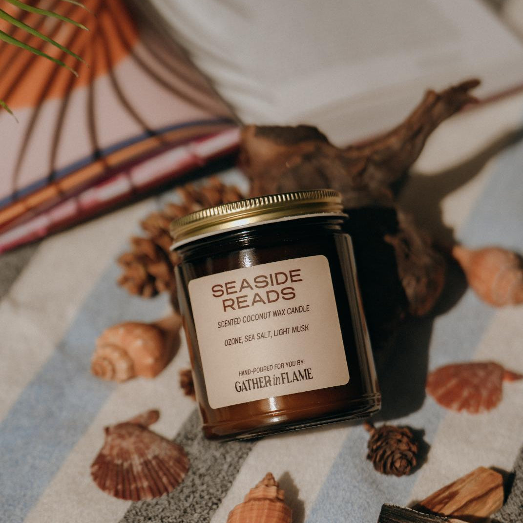 Amber glass candle jar labeled Seaside Reads on a towel surrounded by shells, driftwood and an open book.