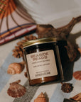 Amber glass candle jar labeled Seaside Reads on a towel surrounded by shells, driftwood and an open book.