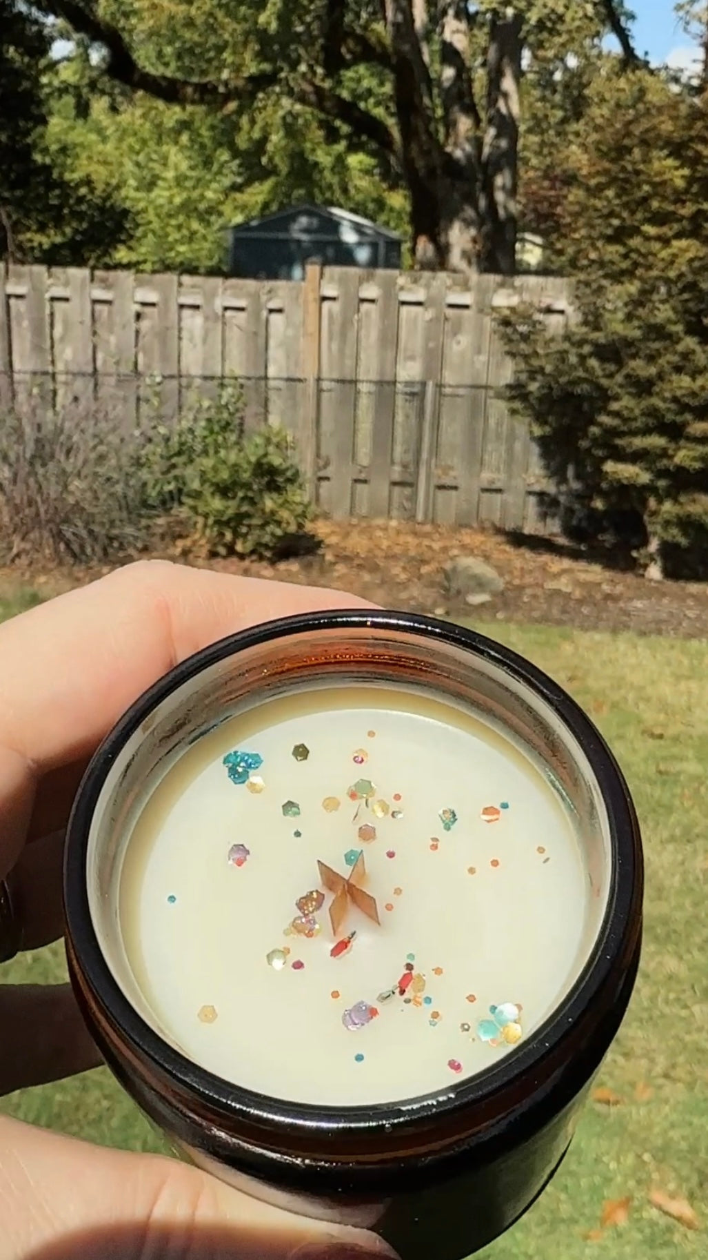 Hand holding a swiftie-inspired candle with colorful glitter sprinkles against a backyard background