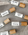 Flatlay photo of six perfume oils with clear glass body and bamboo cap on a tan fabric surface.