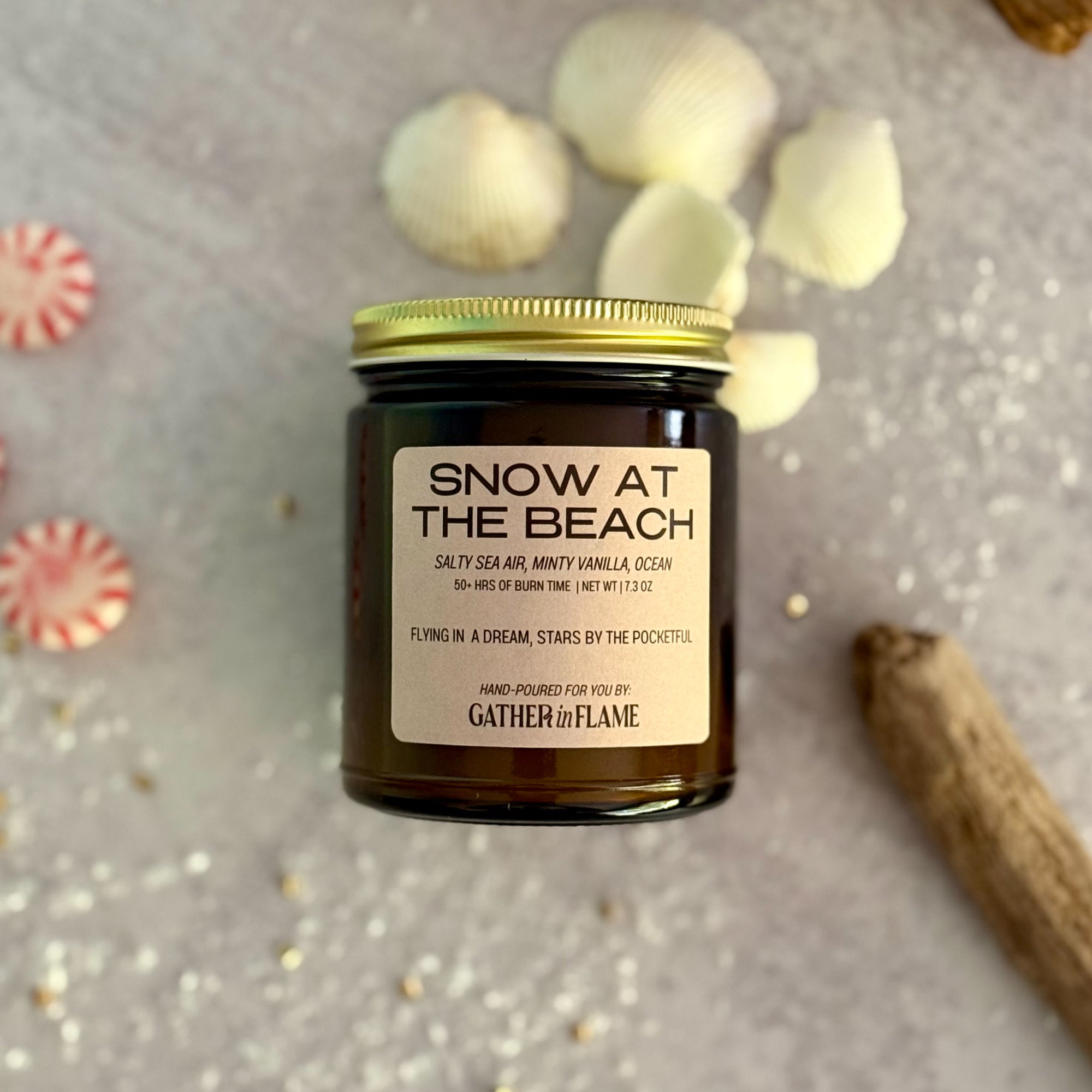 Flatlay showing candle labeled 'Snow at the Beach' by Gather 'n Flame on a textured surface with decorative elements.