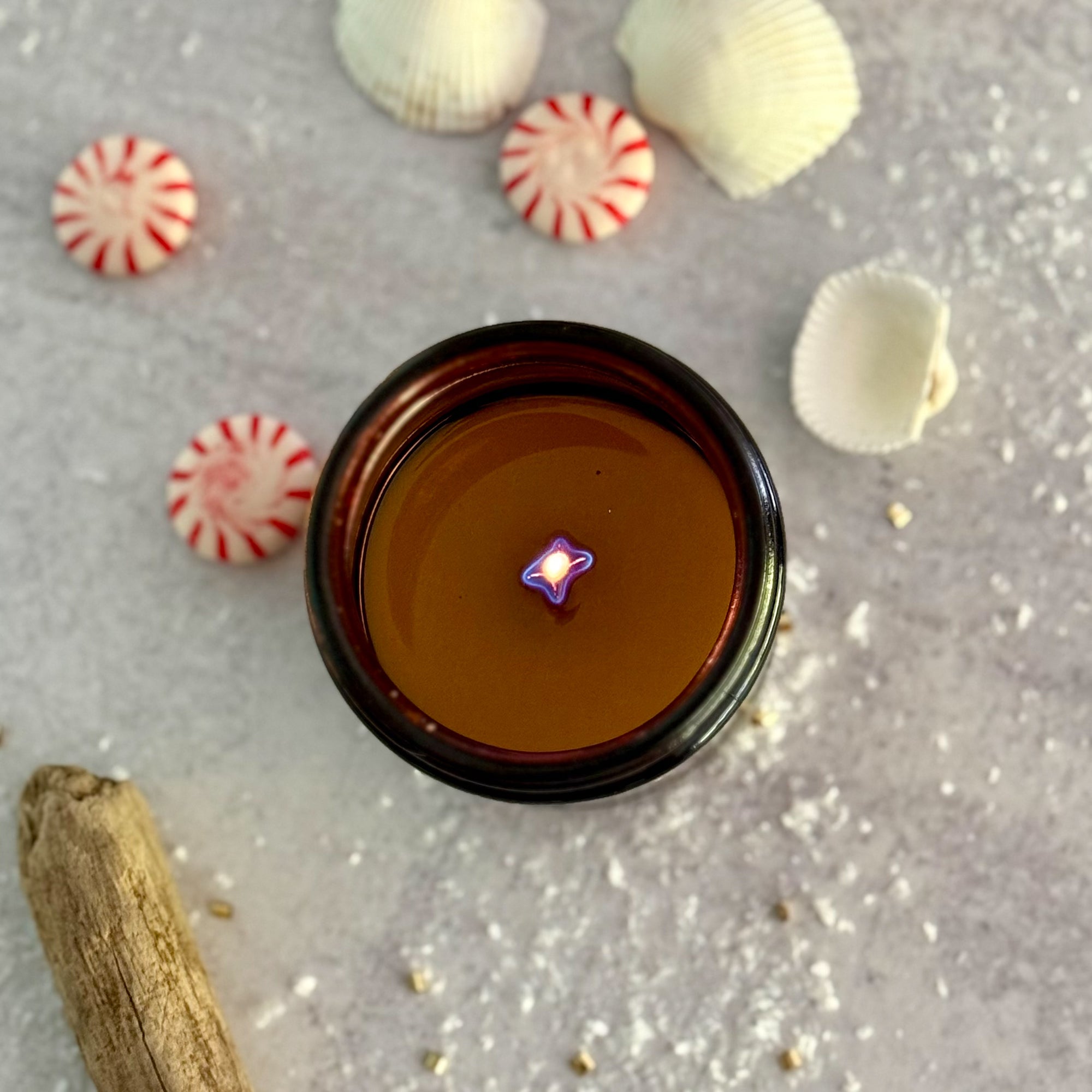 overhead view of lit candle surface with melted wax surrounded by peppermint candies and cinnamon sticks on a gray surface