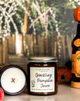 A jar candle with a label reading 'Sparkling Pumpkin Juice' on a tan surface next to a candle showing the smooth surface and wooden x wick with pumpkins and a juice bottle in the background.