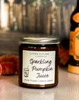 A jar candle with a label reading 'Sparkling Pumpkin Juice' on a tan surface with pumpkins and a juice bottle in the background.