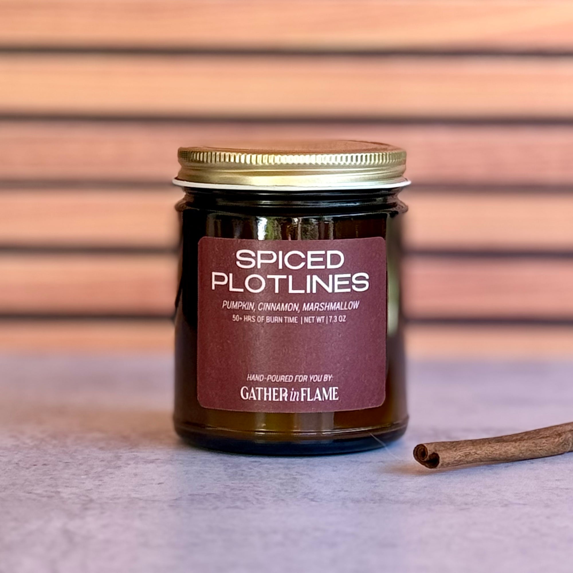 Candle jar labeled 'Spiced Plotlines' with a cinnamon stick on a wooden surface