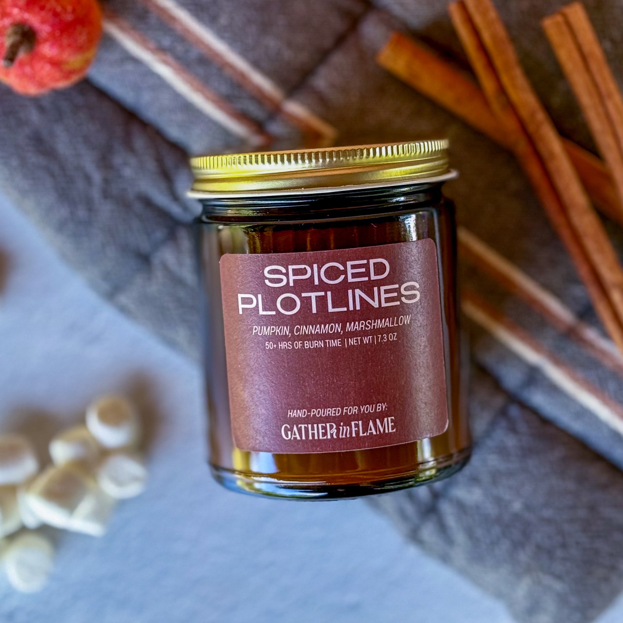 Flatlay photo of 'Spiced Plotlines' candle with cinnamon sticks, mini marshmallows and an small pumpkin in the background