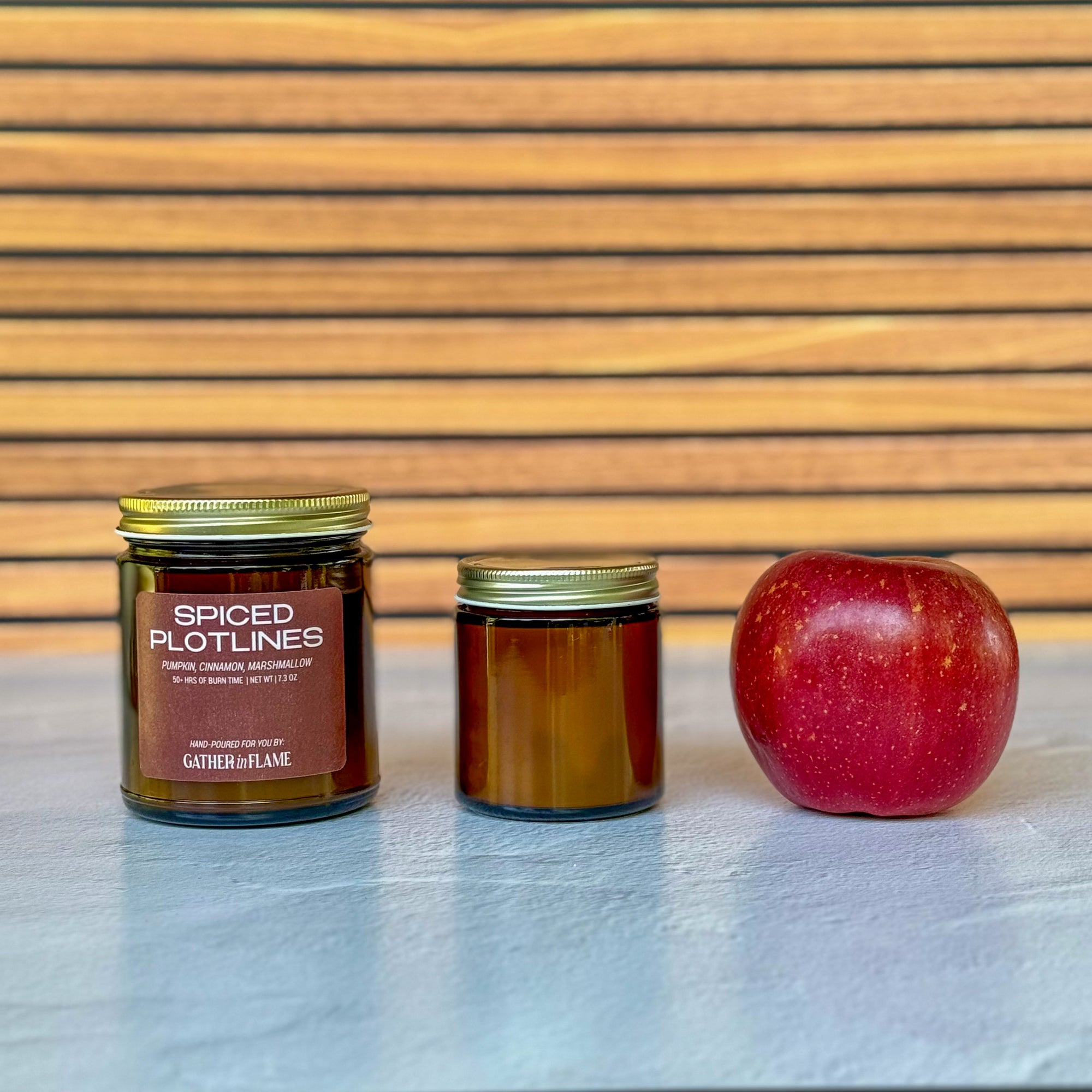 Two candles of different sizes labeled 'Spiced Plotlines' and an apple on a wooden surface with a slatted wood background.